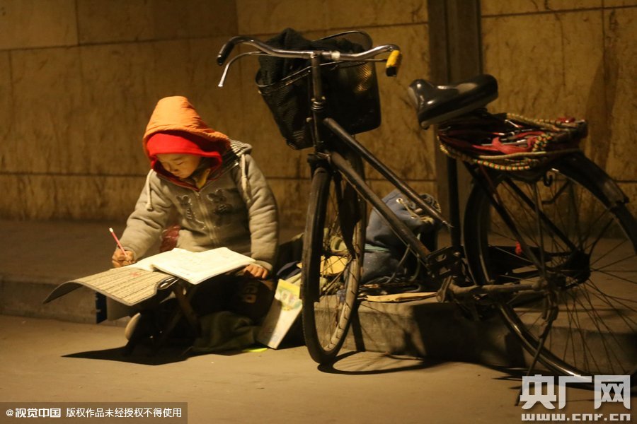 鄭州中原路京廣鐵路大橋下涵洞的馬路牙子邊，一位戴著紅色辣椒帽的小男孩趴在一個(gè)小馬扎上，上面鋪著一張車墊，在微弱的燈光下寫作業(yè)